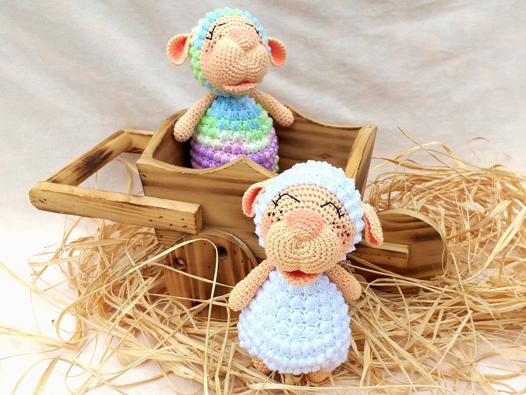 Two pastel crocheted amigurumi sheep on straw, one sitting inside a small wooden wheelbarrow