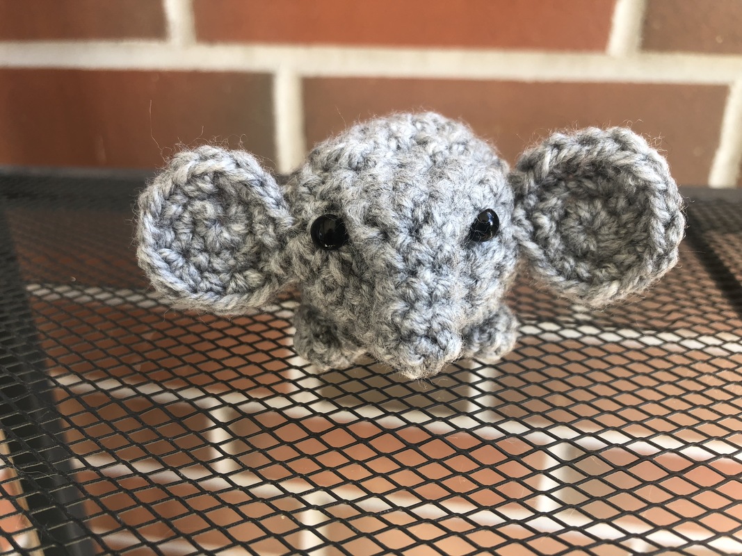 Kleiner grauer Amigurumi-Elefantenkopf mit glänzenden schwarzen Augen auf Metallgitter vor Ziegelwand.