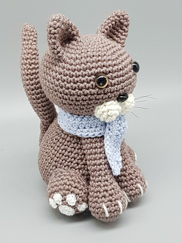 Braune gehäkelte Amigurumi-Katze mit hellblauem Schal und weißen Pfoten vor grauem Hintergrund