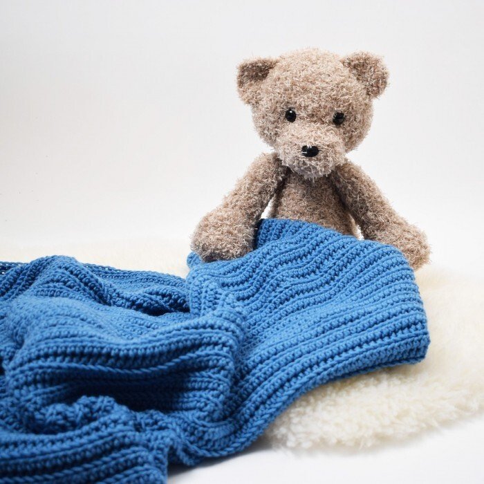 Gehäkelte blaue Babydecke mit welligem Rippenmuster und braunem Teddybär auf weißem Untergrund