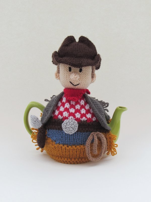 Knitted tea cozy shaped like a cowboy with a brown hat, red bandana and patterned top covering a green teapot.