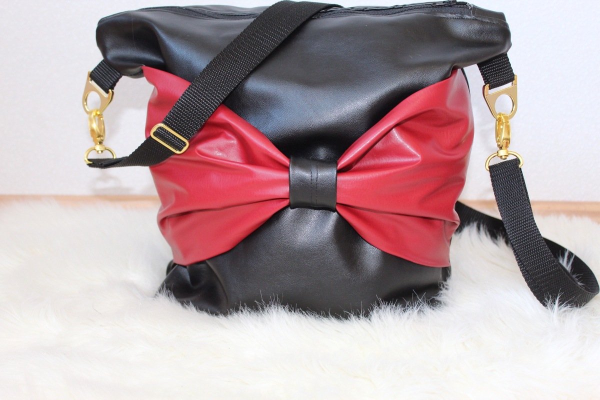 Elegante zweifarbige Umhängetasche in Schwarz und Rot mit großer roten Schleife und goldfarbenen Karabinern am schwarzen Tragegurt.