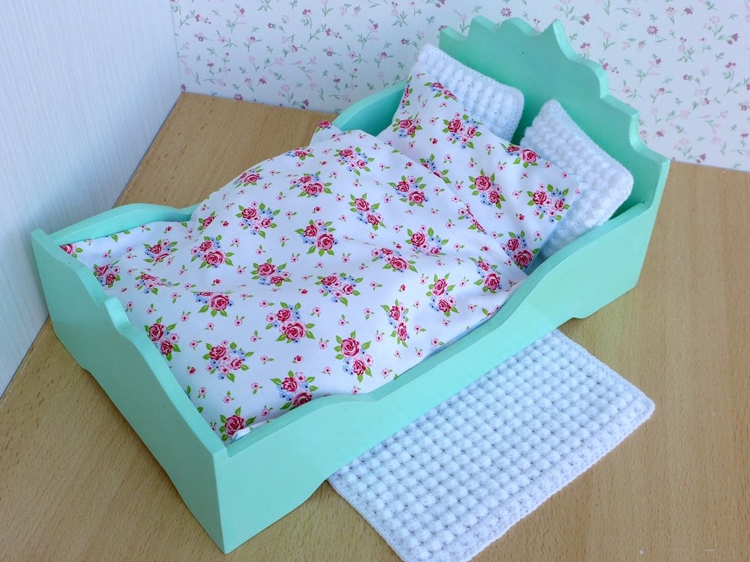 Mintgrünes Puppenbett mit weißer Bettwäsche im Rosenmuster, zwei gehäkelten Kissen und gehäkeltem Teppich auf Holzboden.