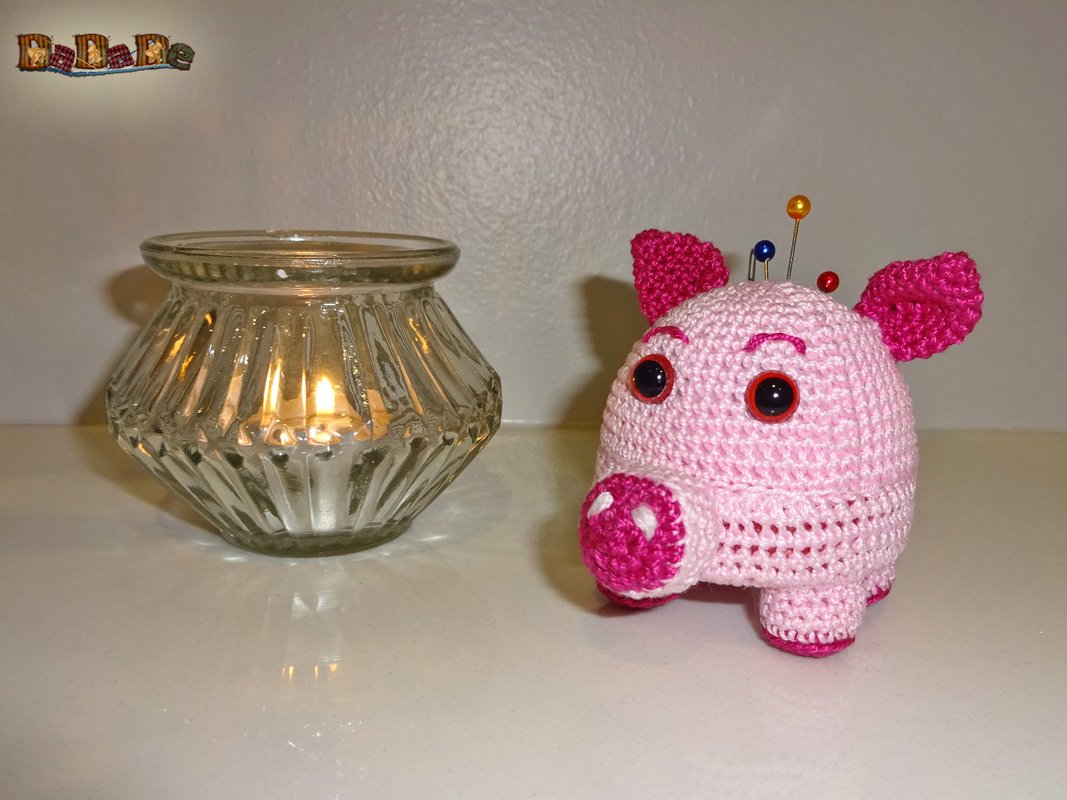 Rosa gehäkeltes Schwein mit Stecknadeln neben brennendem Teelicht in geripptem Glas auf weißer Oberfläche.