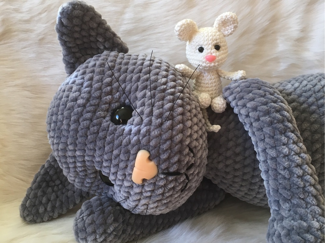 Gehäkelte graue Amigurumi-Katze mit rosa Nase und schwarzen Augen; kleine weiße Amigurumi-Maus sitzt auf ihrem Kopf, auf hellem Fell liegend.