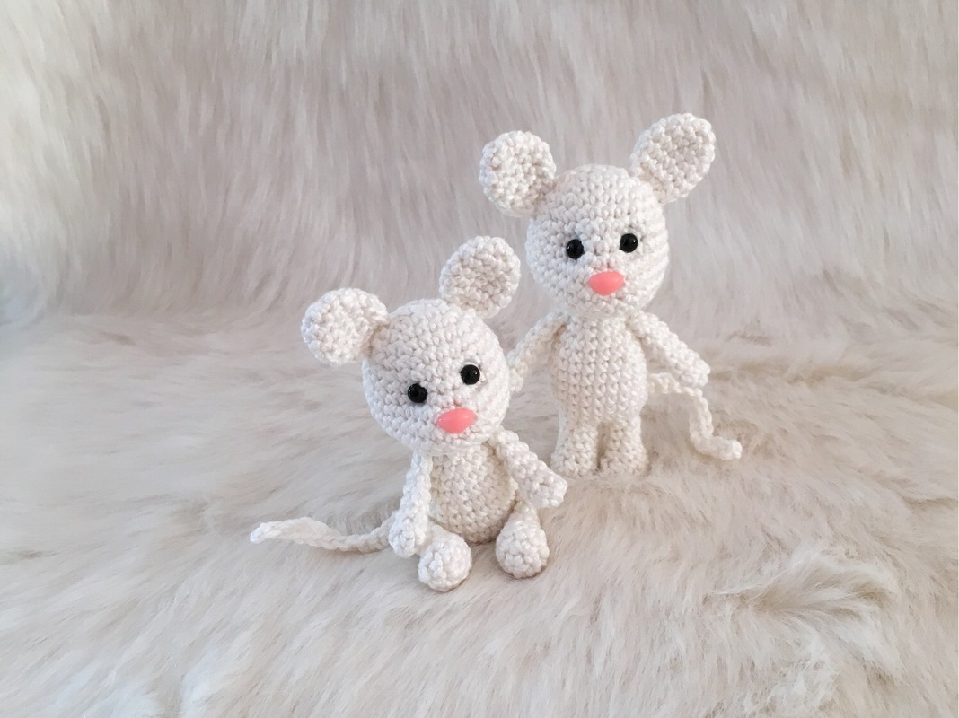 Zwei weiße gehäkelte Amigurumi‑Mäuse mit schwarzen Augen und rosa Nasen auf hellem, flauschigem Hintergrund