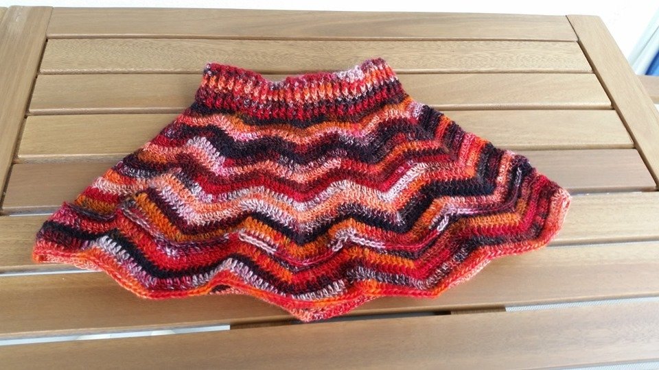 Gehäkelter Kinderrock mit Zickzackmuster in Rot-, Orange-, Schwarz- und Pinktönen auf Holztisch