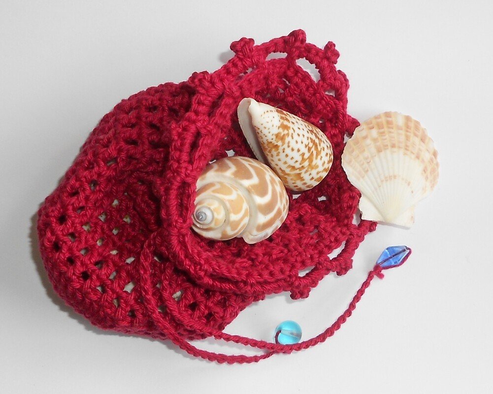Rotes gehäkeltes Säckchen mit drei Muscheln und zwei blauen Perlen auf weißem Hintergrund