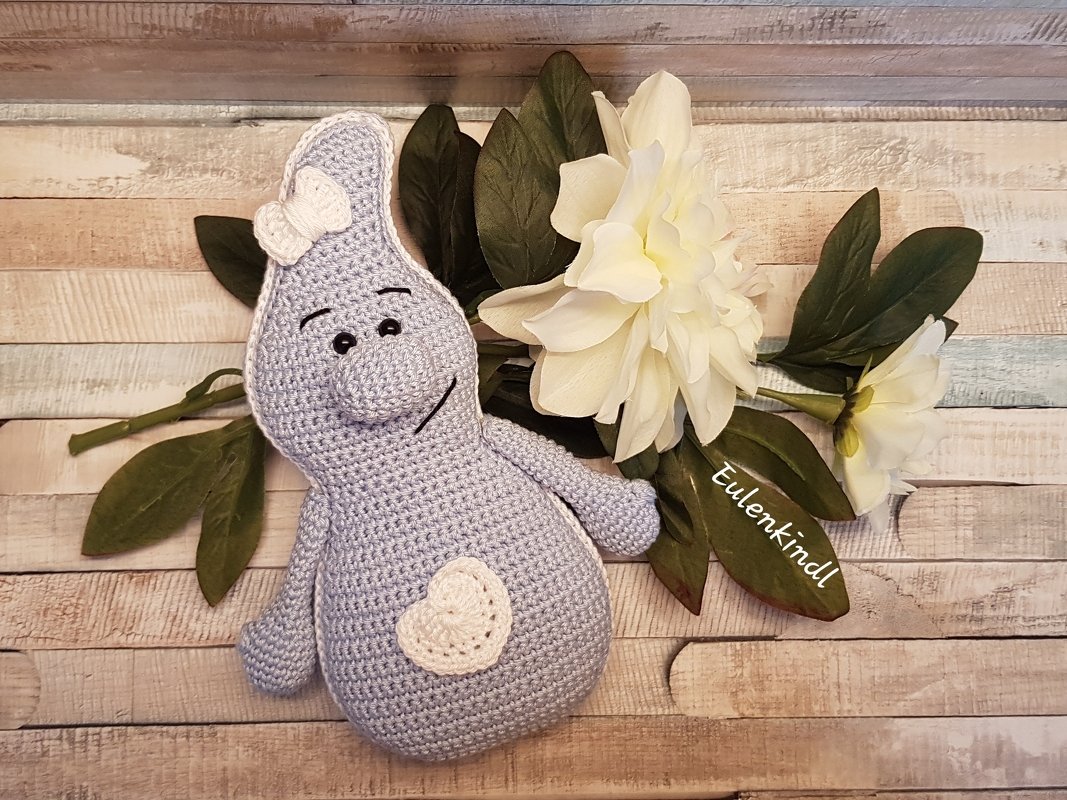 Hellblaues gehäkeltes Amigurumi-Geistchen mit weißer Schleife und Herz vor Holz und Blumen