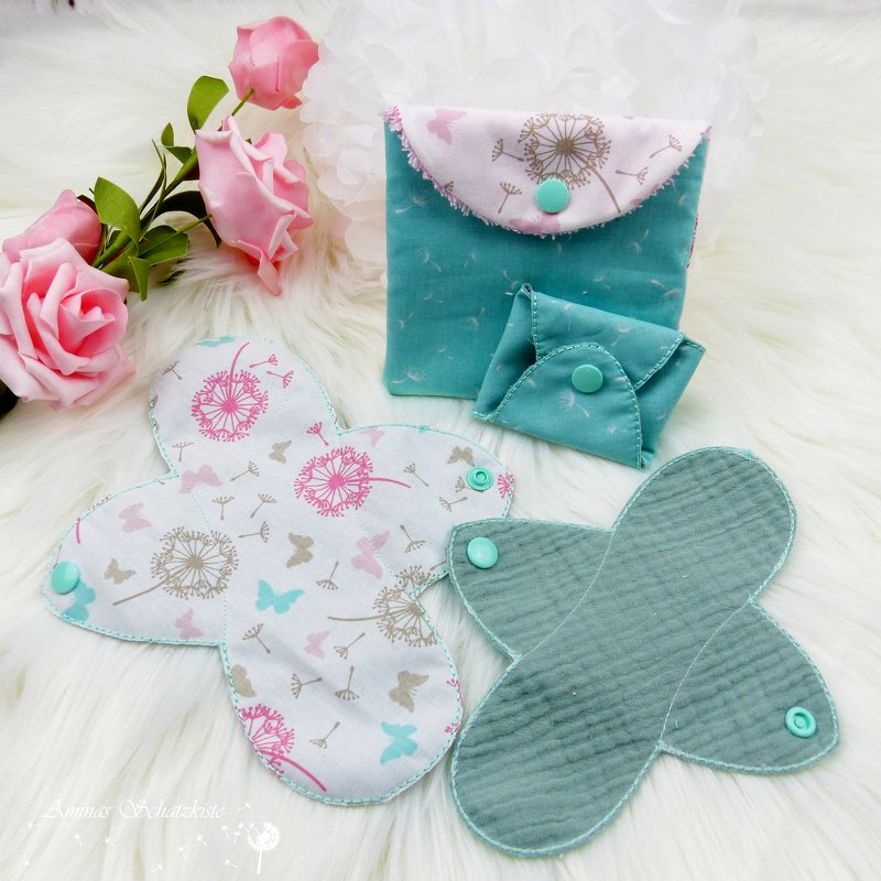 Gemusterte Stoffbinden und zwei Täschchen mit Pusteblumen‑ und Schmetterlingsmotiven in Rosa und Türkis auf weißem Fell.