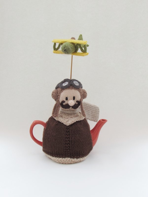 Knitted teapot cozy shaped like a pilot with goggles, mustache and scarf, topped by a small knitted biplane on a wire, sitting on a red teapot