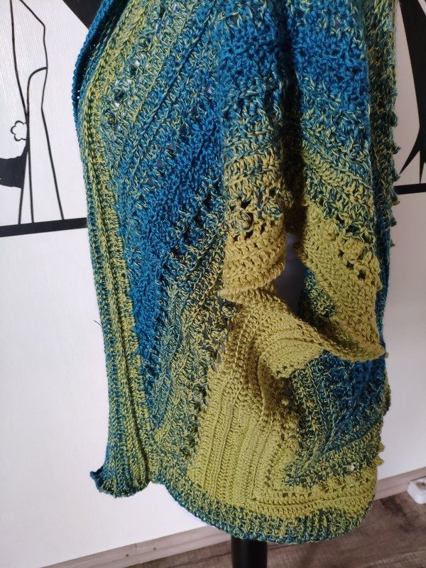 Seitliche Nahaufnahme eines gehäkelten Seelenwärmers in Blau‑Grün mit sichtbaren Strukturmaschen