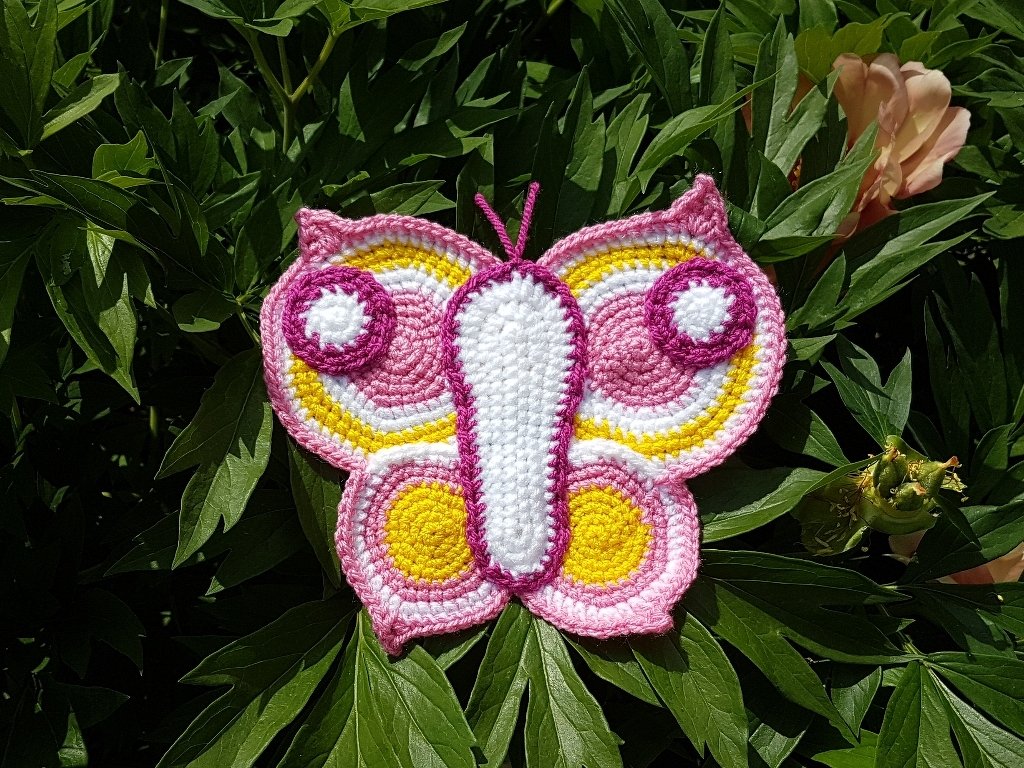 Gehäkelte Schmetterlings-Applikation in Rosa, Gelb und Weiß auf grünen Blättern