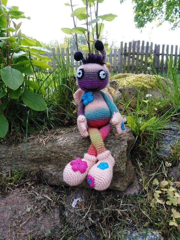 Buntes handgehäkeltes Amigurumi-Insekt mit großen Augen, Antennen und Blumenverzierung auf einem Stein im Garten