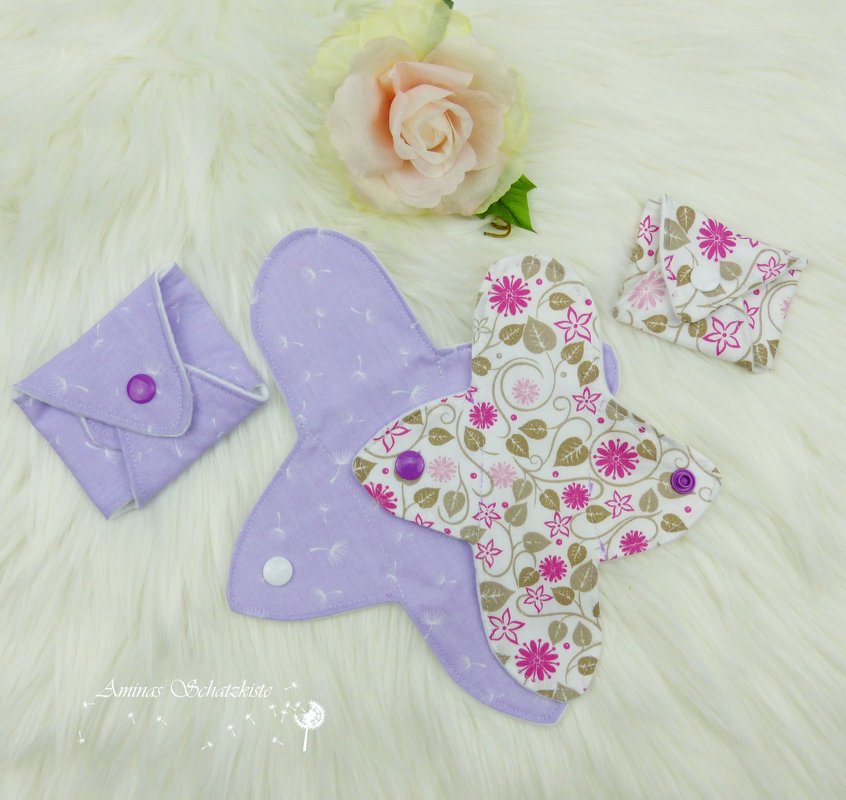 Genähtes Stofftäschchen und textiles Pad mit Flügeln, gefertigt aus lila und floral gemustertem Stoff, arrangiert auf weißem, flauschigem Hintergrund.