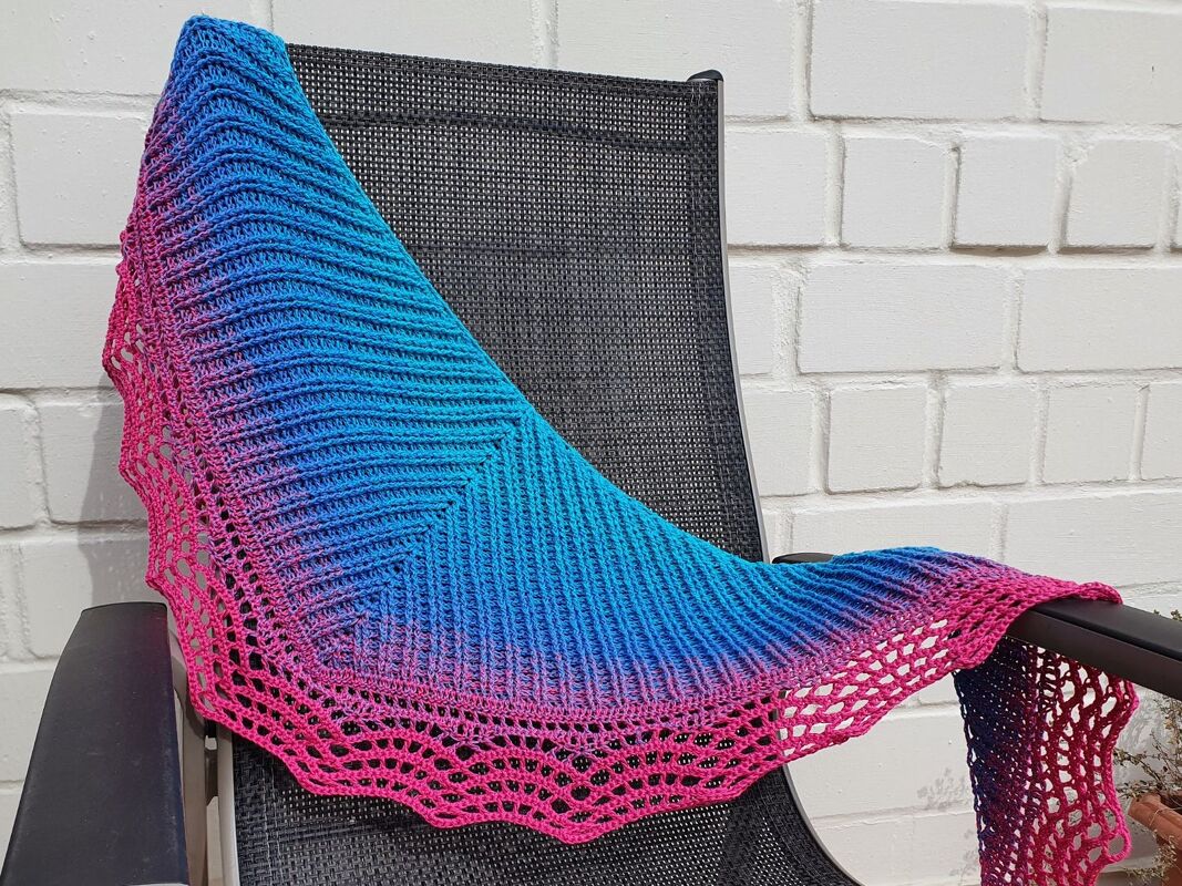 Gehäkeltes Dreieckstuch in Blau und Pink mit Rippenmuster und Spitzenrand auf dunklem Stuhl vor weißer Wand.