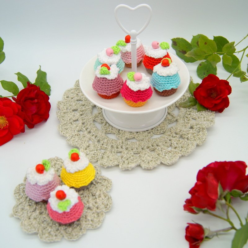 Weiße Etagere mit bunten gehäkelten Mini-Törtchen; zusätzliche Törtchen auf einem gehäkelten Deckchen und rote Rosen als Dekoration.