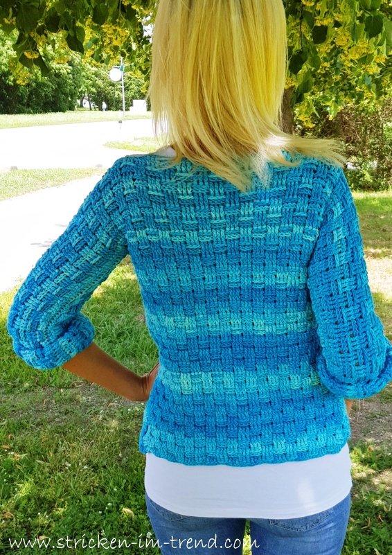 Back view of a woman wearing a blue and turquoise crocheted jacket with a basketweave-like textured stitch and three-quarter sleeves, standing outdoors.