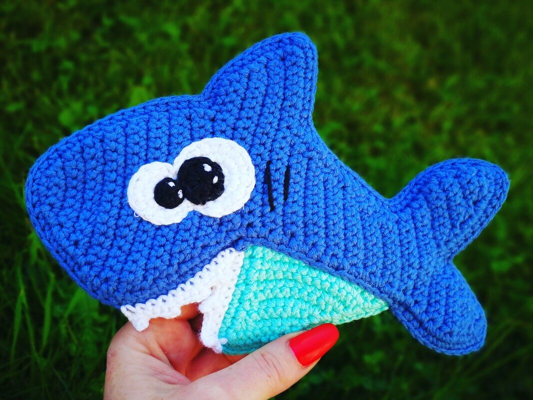 Handgehäkeltes blaues Haifisch-Spielzeug mit großen weißen Augen und angedeuteten Zähnen, in einer Hand vor grünem Gras gehalten.