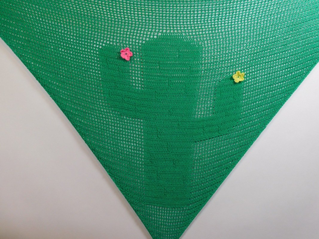 Grünes gehäkeltes Dreieckstuch mit eingearbeitetem Kaktusmotiv und zwei kleinen Häkelblumen in Rosa und Gelb.