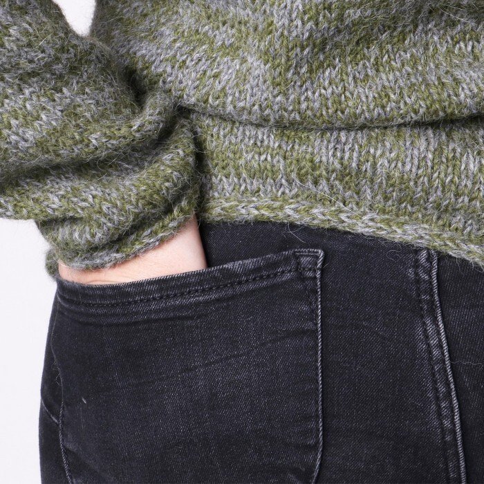 Close-up of a woman wearing a green and grey knitted sweater with a hand in a black jeans pocket.