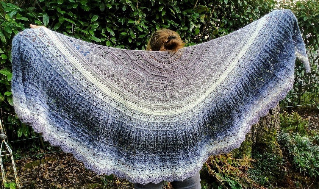 Person hält großes halbrundes gehäkeltes Tuch mit Farbverlauf in Grau, Flieder und Creme, gewellte Spitzenkante sichtbar