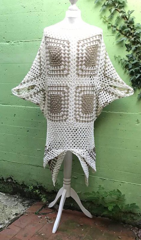 Gehäkeltes Granny‑Square‑Kleid in Creme und Hellbraun auf einer Schneiderpuppe vor grüner Wand mit Efeu; offene Maschen und weiter Schnitt sichtbar.