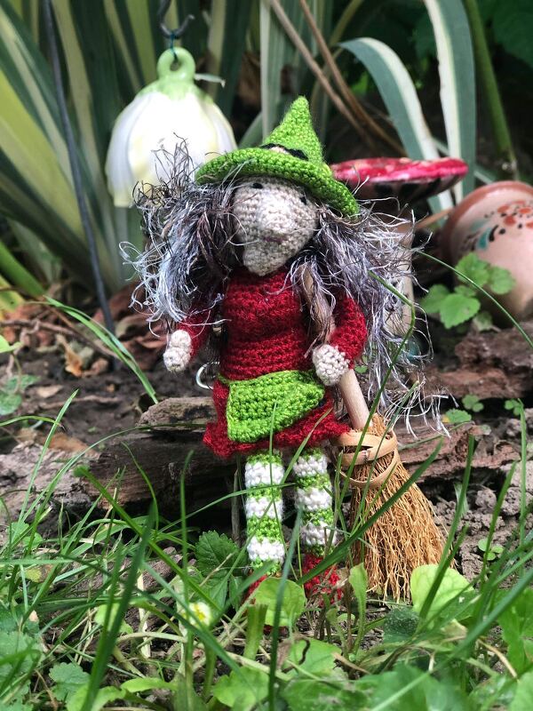 Gehäkelte Hexenpuppe mit grünem Hut, rotem Kleid und Besen im Gras