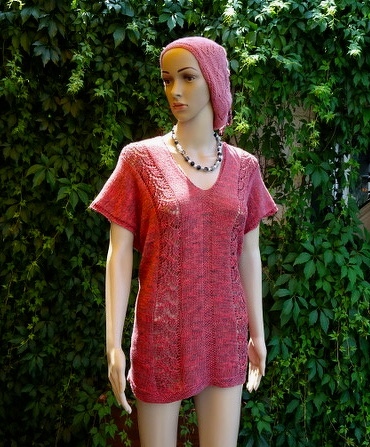 Mannequin trägt rosarote gestrickte Tunika mit V-Ausschnitt, Kopftuch und Perlenkette vor grünem Hintergrund