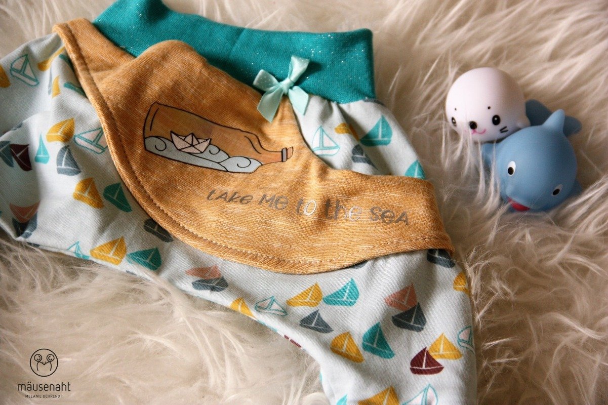 Babystoff mit kleinem buntem Segelboot-Muster, senffarbene Klappe mit Flaschenmotiv und Papierboot, zwei Spielzeugtiere auf Felluntergrund, kleines rundes Logo in Ecke