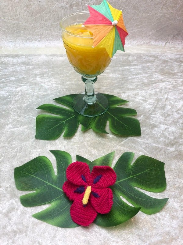 Gehäkelte rote Hibiskusblüte mit gelbem Stempel und violetten Akzenten auf grünen, tropischen Blättern; im Hintergrund ein Glas mit orangefarbenem Getränk und buntem Papier-Sonnenschirm.