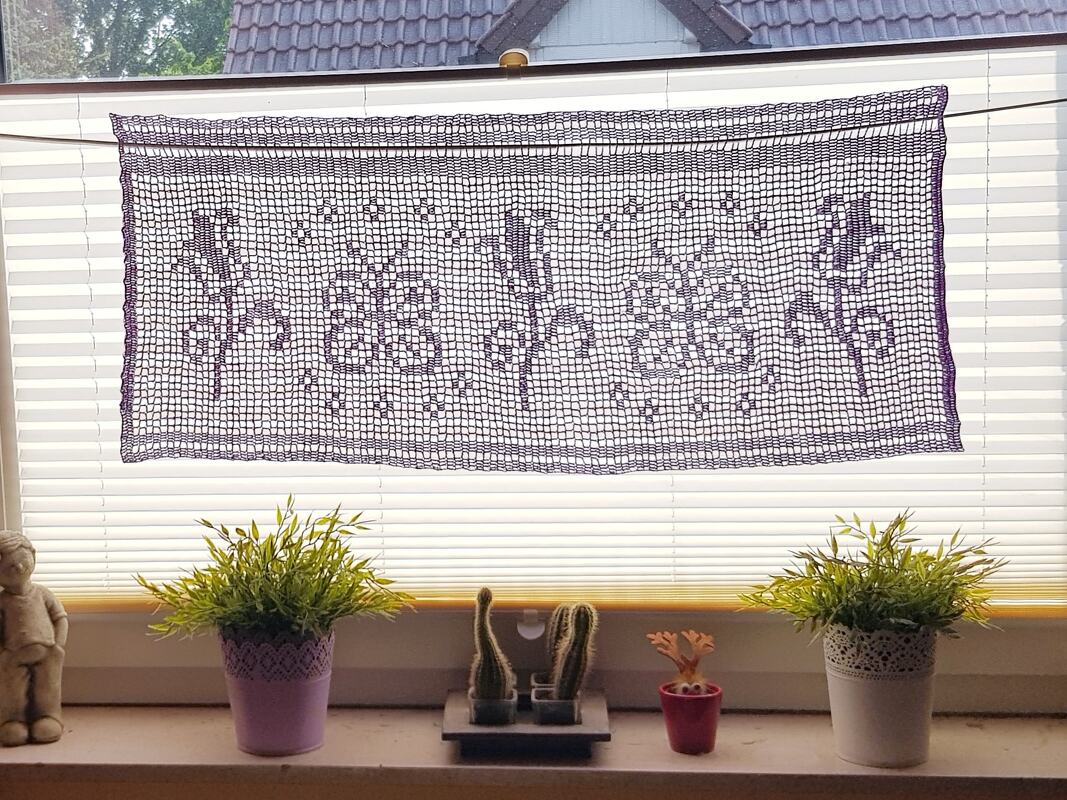 Dunkelviolette gehäkelte Filetgardine mit Blumen- und Rankenmuster vor einem Fenster, darunter Pflanzen auf der Fensterbank.