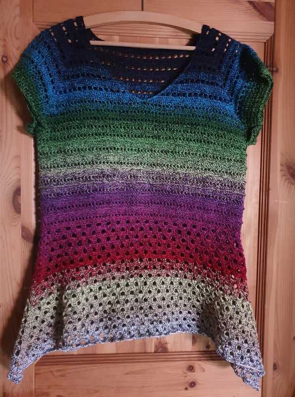 Gehäkeltes Kurzarm-Shirt mit Farbverlauf und Lochmuster an Holzbügel vor einer Holztür