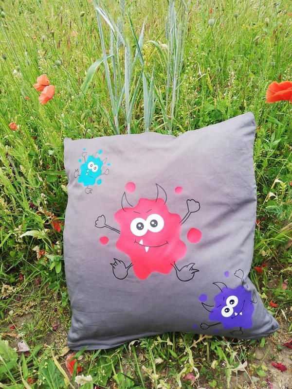 Graues Kissen mit drei bunten Klecks‑Monstern in Rot, Lila und Blau auf einer Wiese