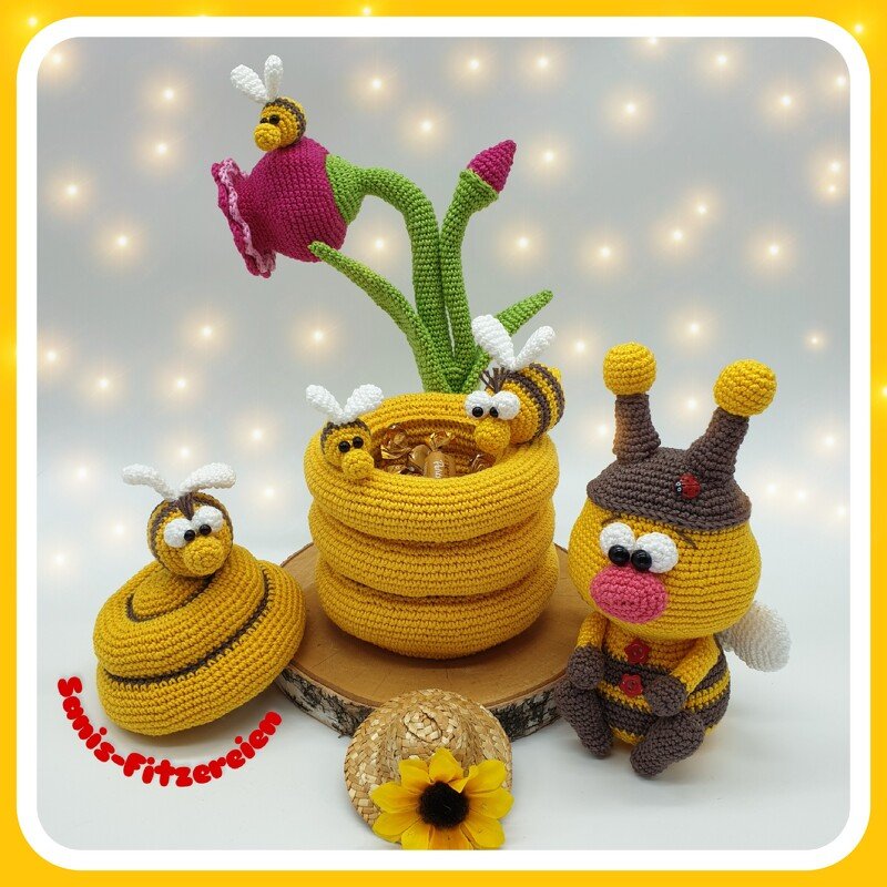 Mehrere gehäkelte Bienen-Amigurumis, ein gelber bienenkorbförmiger Korb, eine rosa Blüte und eine gelb-braun gestreifte Bienenfigur mit Fühlern auf hellem Hintergrund