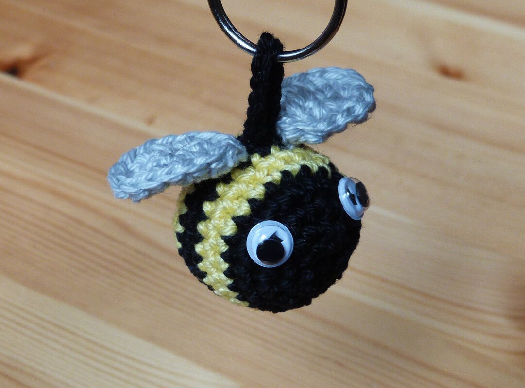 Gehäkelter Bienen-Schlüsselanhänger mit schwarz-gelben Streifen, grauen Flügeln und Wackelaugen an einem Schlüsselring vor Holz.