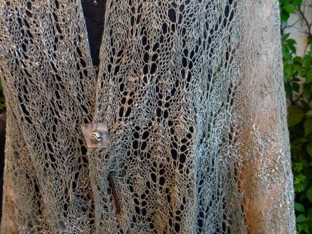 Nahaufnahme eines hellgrauen Lacetuchs mit offenem Lochmuster und quadratischer Brosche über dunkler Kleidung