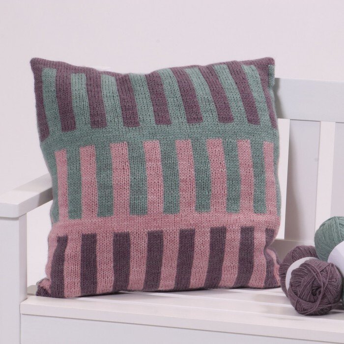 Knitted pillow with vertical block stripes in mauve, pink and green beside yarn balls