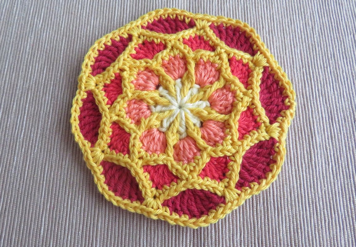 Runder gehäkelter Untersetzer mit gelbem Gitter, rosa und korallfarbenen Segmenten und weißem Zentrum auf hellem Stoff