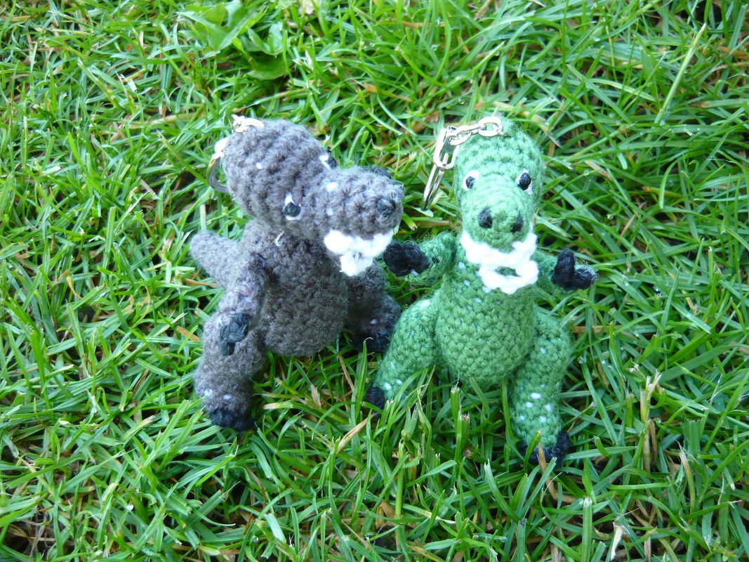 Zwei gehäkelte Dinosaurier‑Schlüsselanhänger, grün und grau, auf Gras
