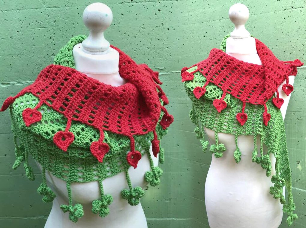 Grüner gehäkelter Dreiecksschal mit roter Randblende, roten Herzanhängern und grünen Kleeblattfransen, an einer Schneiderpuppe drapiert