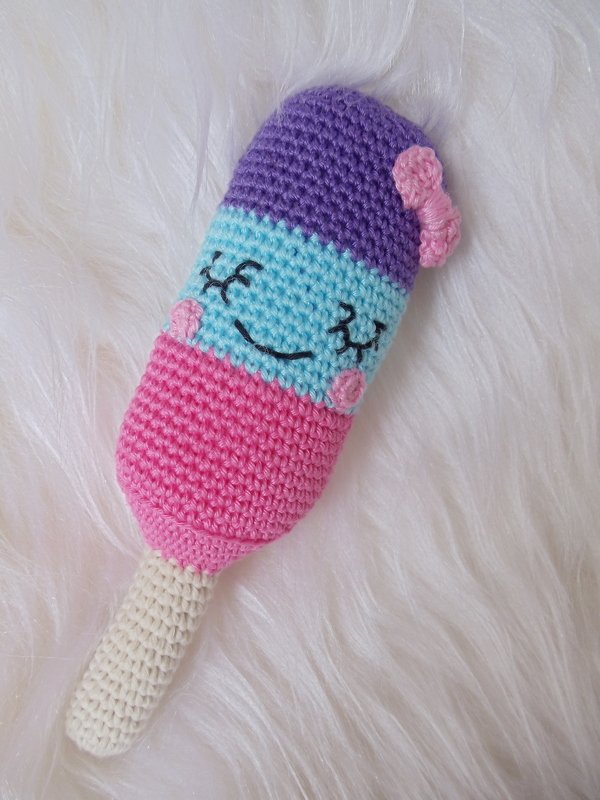 Gehäkeltes Eis am Stiel Amigurumi in Violett, Hellblau und Pink mit gesticktem Gesicht und Schleife auf weißem, flauschigem Untergrund.