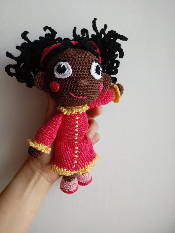 Crocheted amigurumi doll with dark brown complexion, black curly hair, pink dress and pink shoes held in a hand.