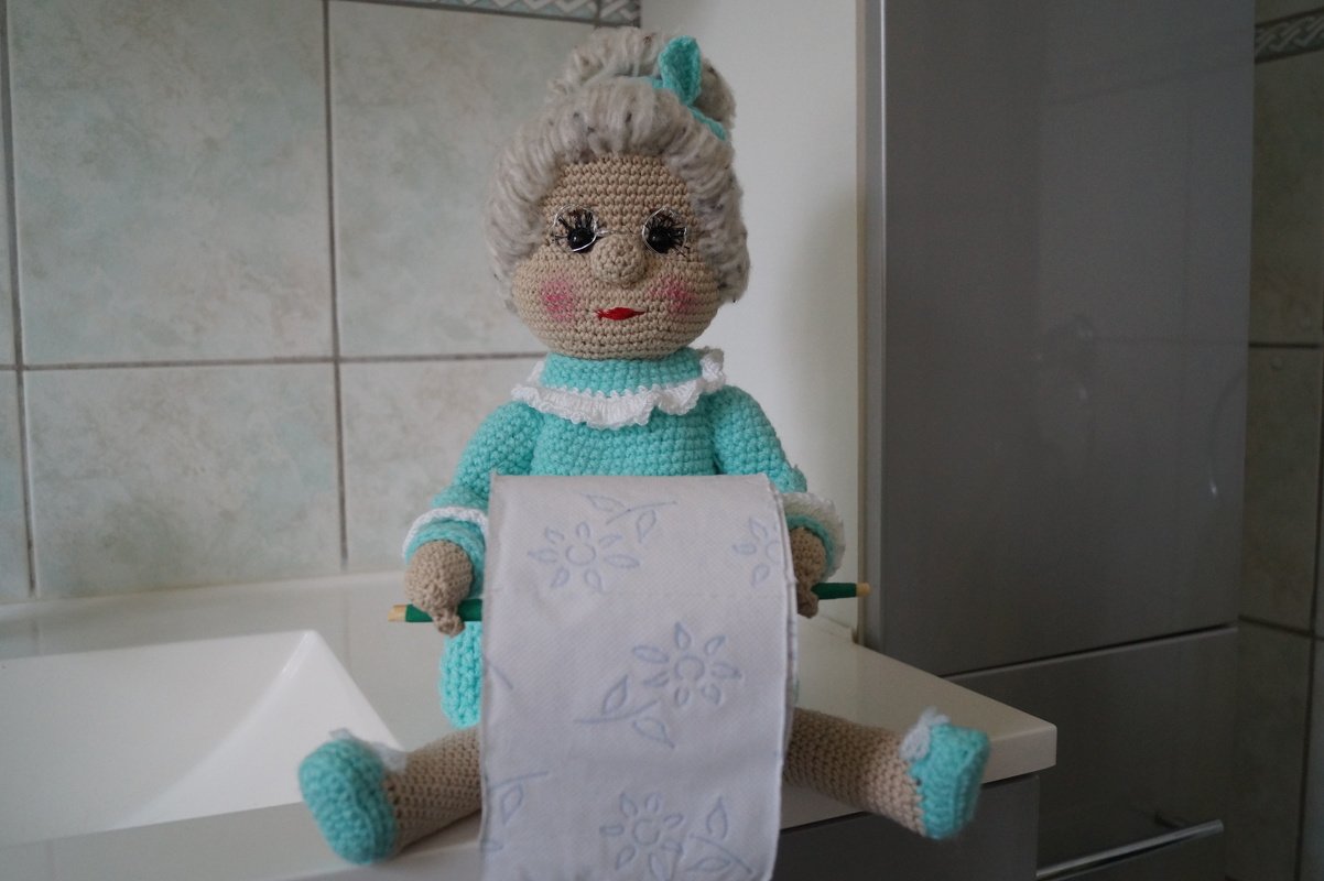 Gehäkelte Oma-Puppe mit grauem Haar und türkisfarbenem Kleid hält eine Toilettenpapierrolle auf einer Ablage.