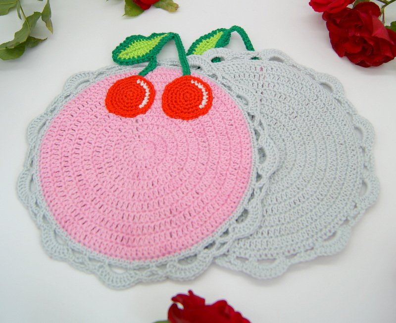 Zwei runde, gehäkelte Topflappen auf hellem Hintergrund: einer rosa mit Kirsch-Applikationen, der andere hellgrau.