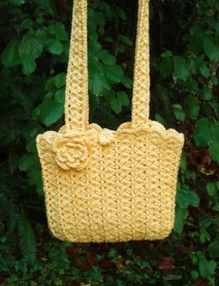 Pale yellow crocheted shoulder bag with flower appliqué hanging against green foliage