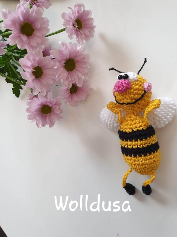Gehäkelte Amigurumi-Biene in Gelb und Schwarz mit weißen Flügeln neben rosa Blüten