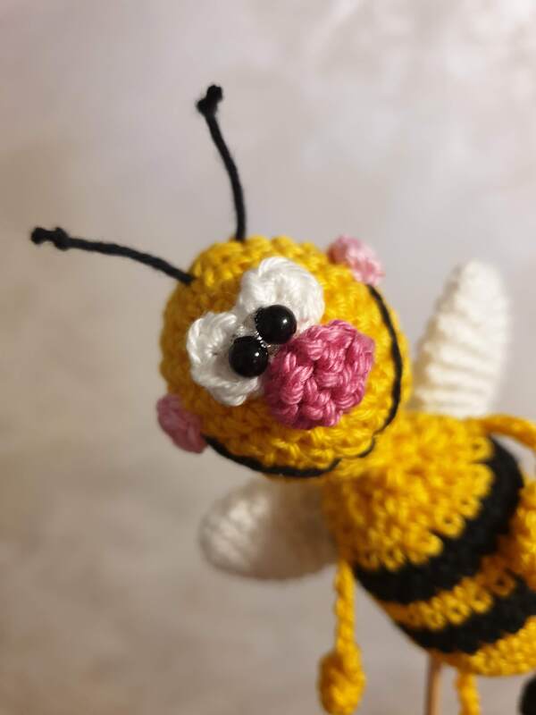 Nahaufnahme einer gelb‑schwarzen gehäkelten Amigurumi‑Biene mit weißen Flügeln, rosa Wangen und schwarzen Perlenaugen