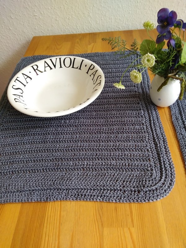 Gehäkeltes anthrazitfarbenes Tischset auf Holztisch mit weißer Schale und Blumen