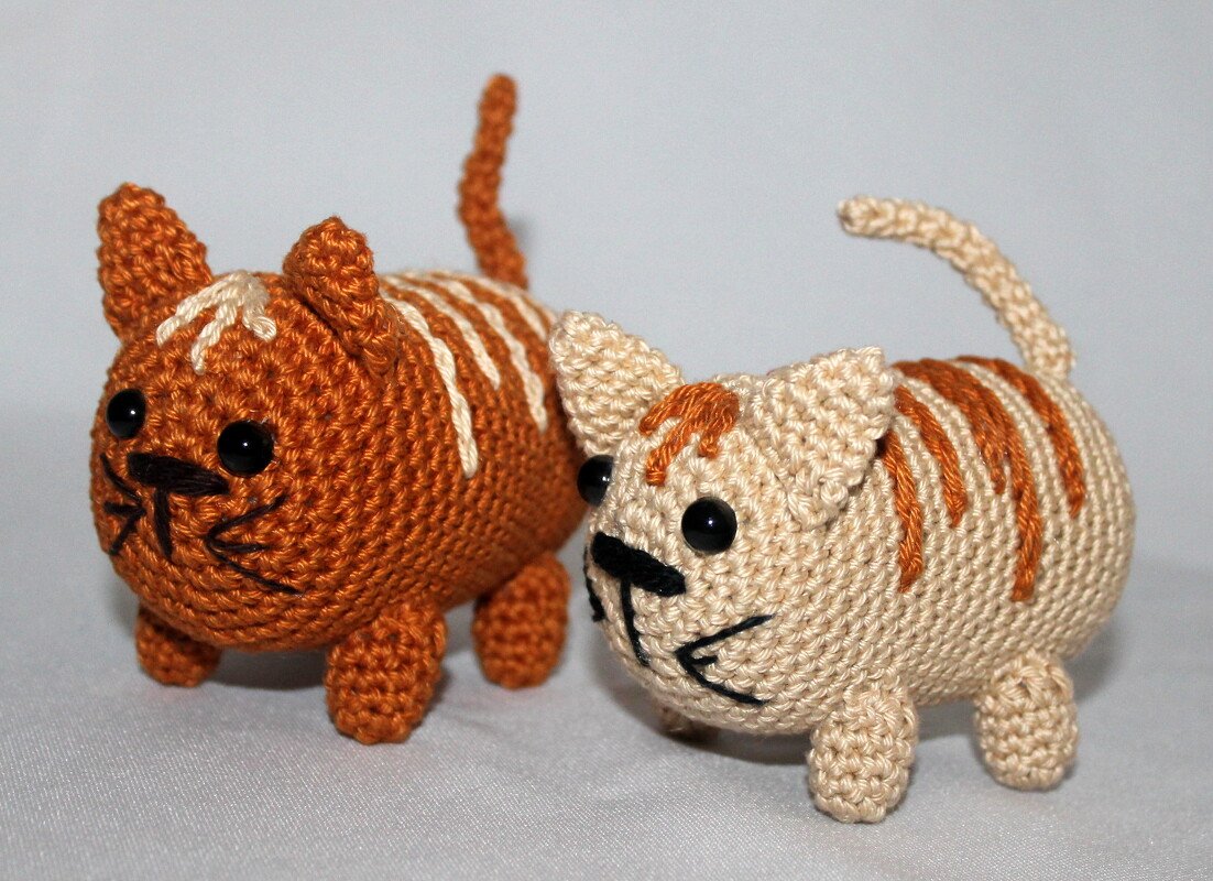 Zwei gehäkelte Amigurumi-Katzen, eine orange mit hellen Streifen, die andere beige mit orangefarbenen Streifen, auf hellem Hintergrund.